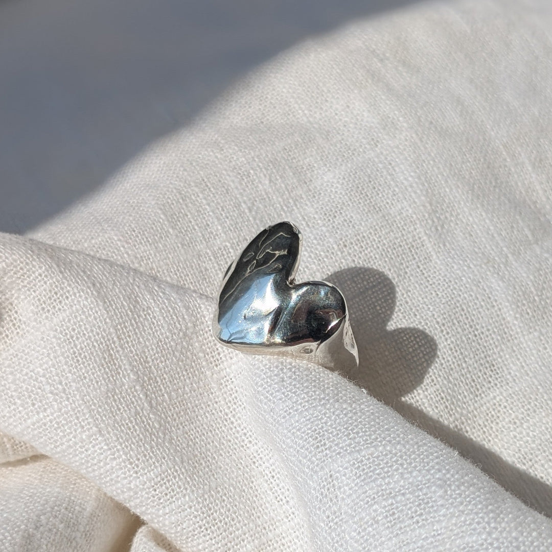 The Gra Heart Ring by Becca Calmont, a handcrafted heart-shaped piece, shines on off-white fabric and glows beautifully in natural sunlight.