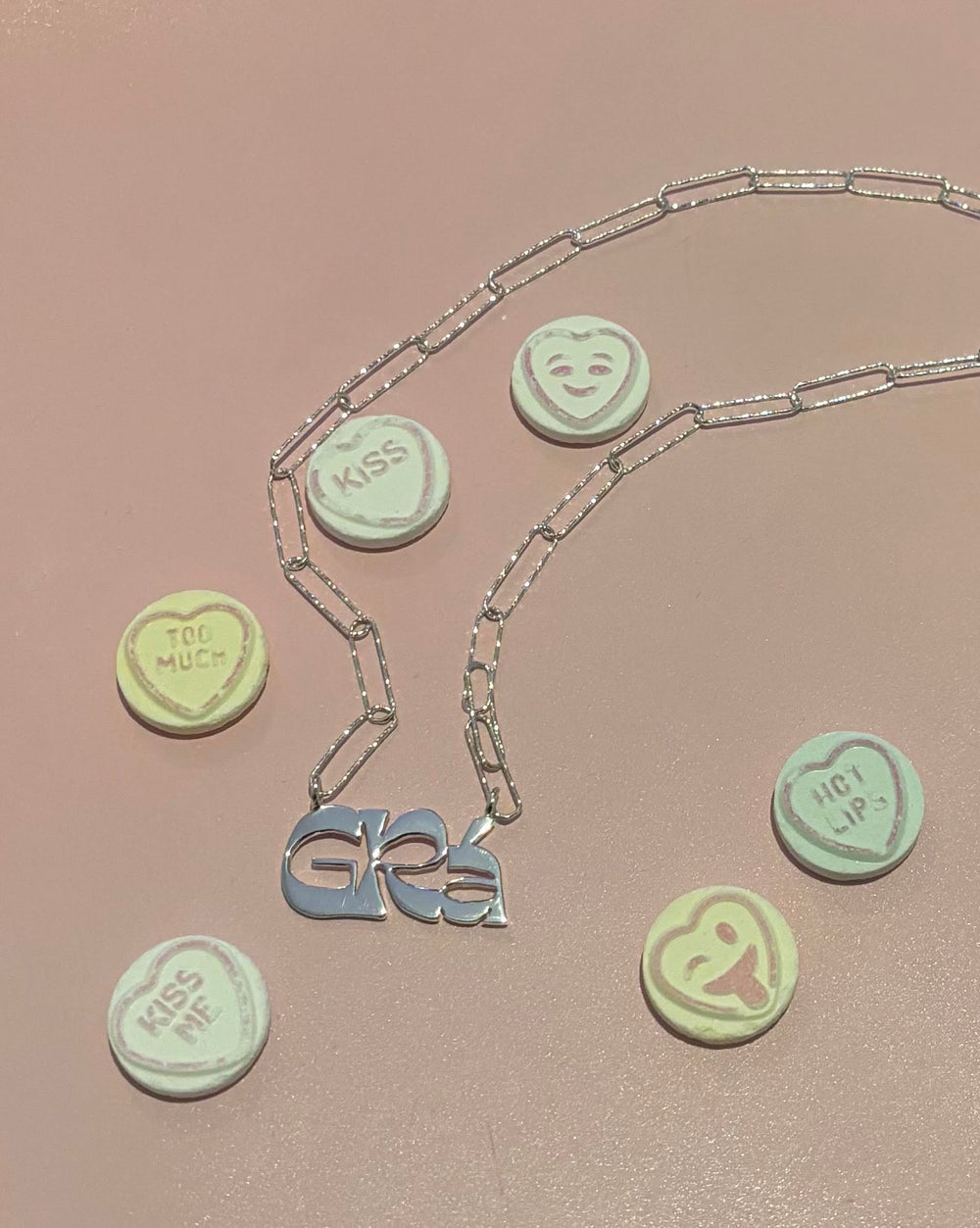 The Cosmic Boulevard Grá Silver Pendant, featuring a "GRL" design, is displayed on a pink surface surrounded by pastel candy hearts with phrases like "TOO MUCH," "KISS ME," and smiley faces.