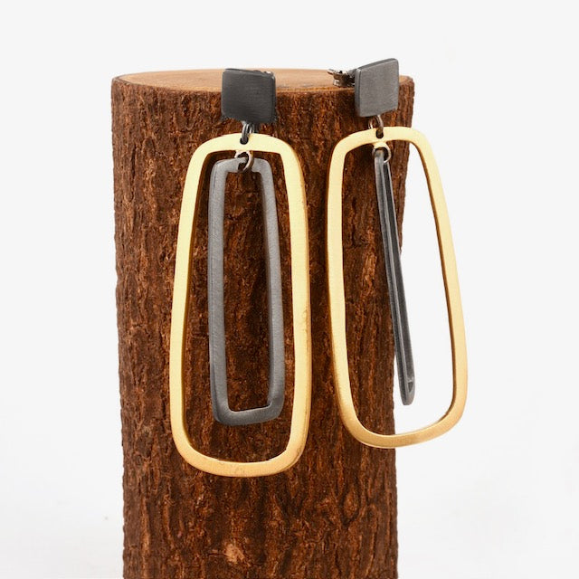 A pair of Grafton Black & Gold Earrings by The Collective Dublin displayed on a wooden stand. Each earring consists of a rectangular black segment enclosed within a larger gold rectangle, creating a stylish, contemporary look against a neutral background.