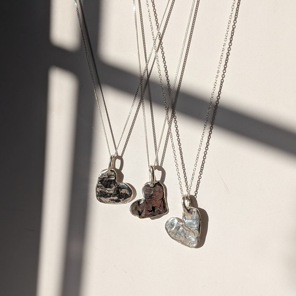 Four Gra Love Heart Pendants by Becca Calmont hang on chains against a light backdrop, sunlight accentuating the textured recycled silver—an ideal display of handcrafted jewelry.