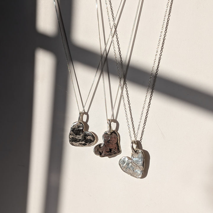 Four Gra Love Heart Pendants by Becca Calmont hang on chains against a light backdrop, sunlight accentuating the textured recycled silver—an ideal display of handcrafted jewelry.
