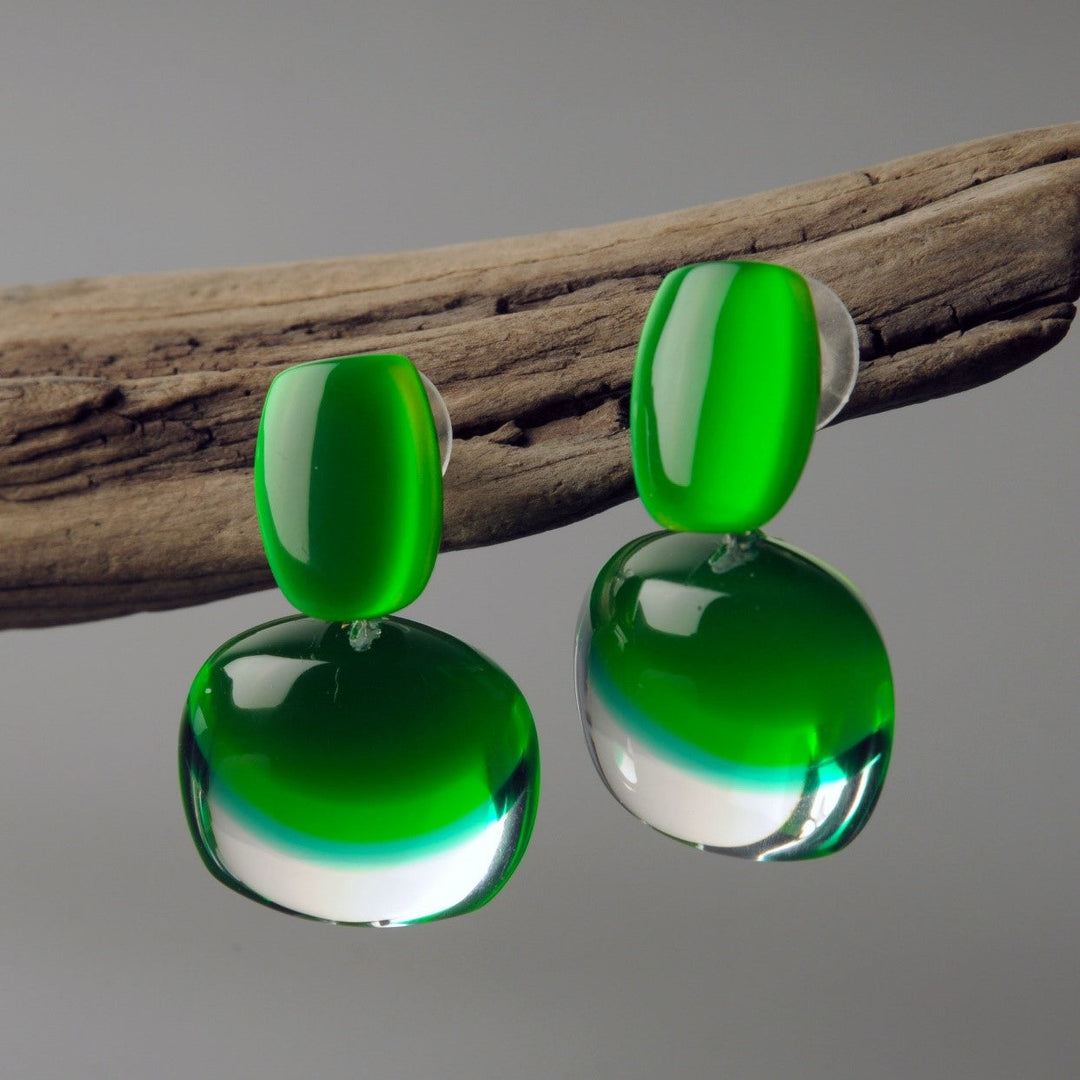 A pair of modern, asymmetrical Acrylic Green Earrings from MX.design displayed on a piece of driftwood. The earrings showcase glossy, emerald green upper sections and clear lower sections with a hint of green, creating a chic, two-toned effect. The background is a plain gray backdrop.