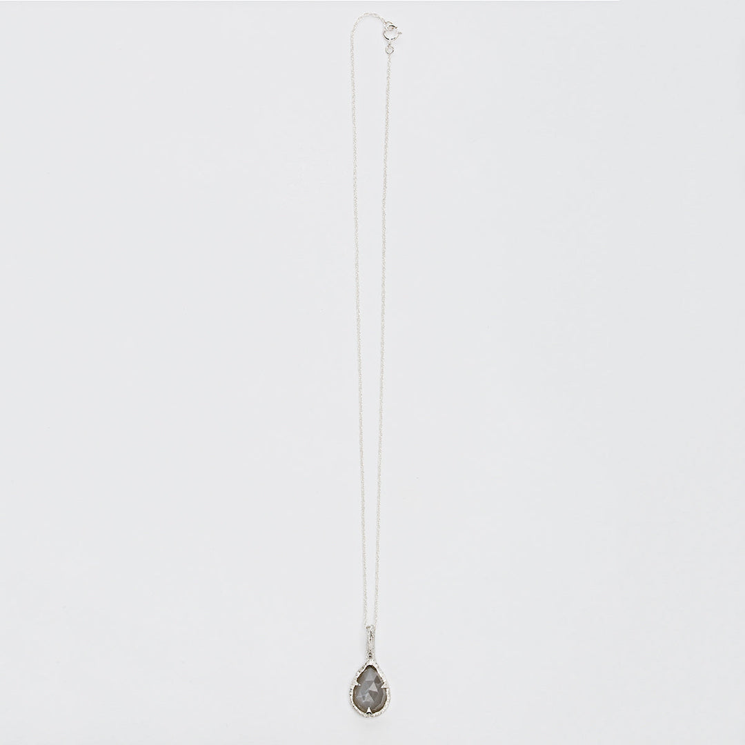 A delicate sterling silver chain necklace with a teardrop-shaped, faceted dark gemstone pendant. The Thorn Teardrop Pendant by Abbie Dixon, which evokes the elegance of a moonstone's glimmer, is displayed against a plain white background.