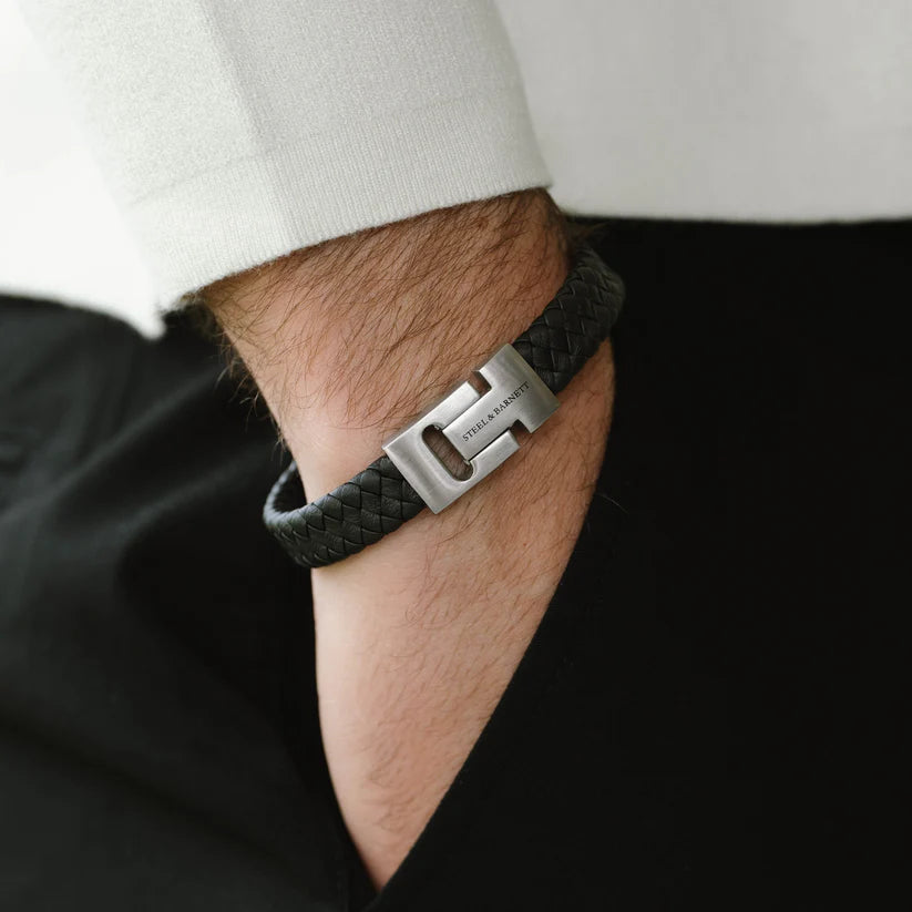 A person wearing a white long-sleeve shirt and black pants has their hand in the pocket. Their wrist sports a sophisticated accessory—a Harrison - Black braided bracelet with a silver clasp from Steel and Barnett, tucked under the cuff of the shirt.