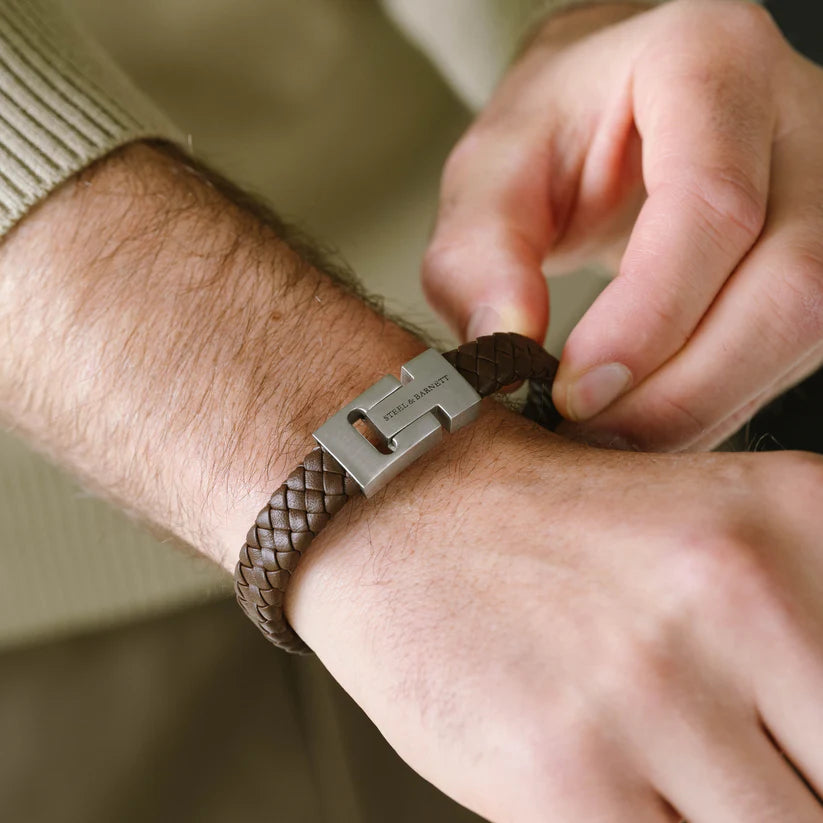 A person wearing a Steel and Barnett Harrison bracelet in brown Nappa leather, featuring a silver clasp, fastened on their wrist. They are dressed in a light-colored, long-sleeved top and adjusting the bracelet with their other hand, exuding the look of a modern professional.