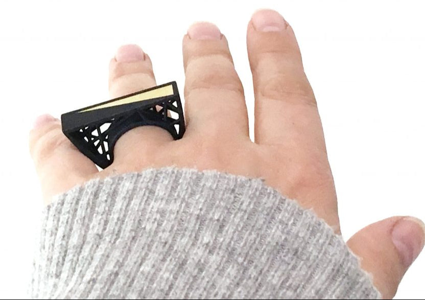 A hand with a beige sleeve wears a large, rectangular-shaped black ring with intricate cut-out detailing and a gold stripe on the top. The Hebo Statement Ring by Elsiem, inspired by Barbara Hepworth's designs, stands out against the plain white background.