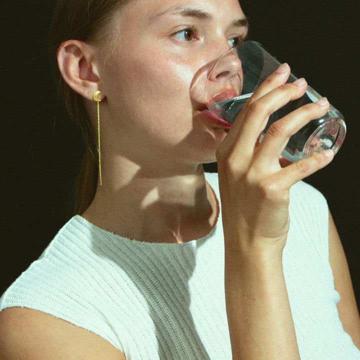 A person with light skin, wearing a sleeveless white top and Jane Koenig’s Half Moon Chain Stud earrings, drinks water from a clear glass against a dark background.