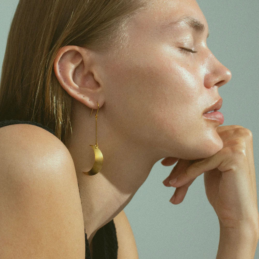 A woman with light skin and straight brown hair, eyes closed, rests her chin on her hand. She wears a sleeveless black top and a striking Jane Koenig Half Moon Earring. The background is light gray.