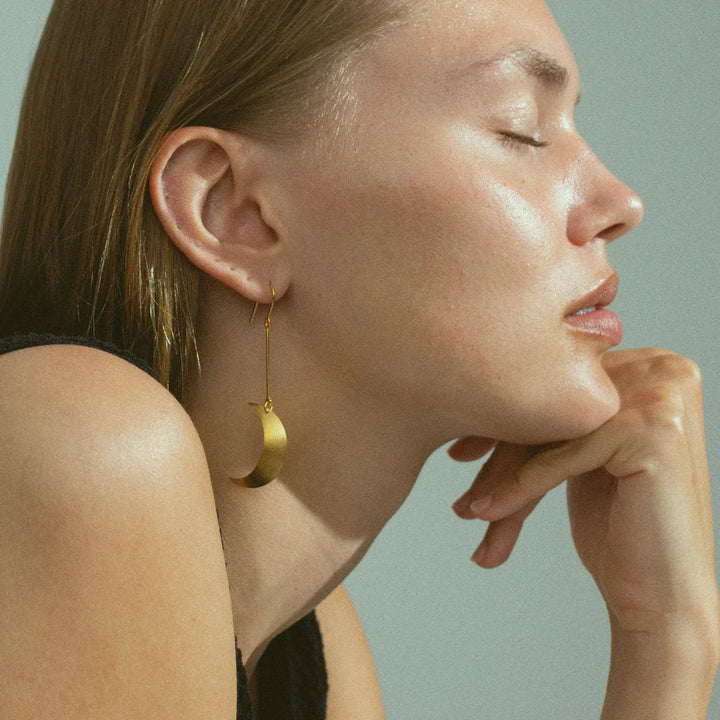 A woman with light skin and straight brown hair, eyes closed, rests her chin on her hand. She wears a sleeveless black top and a striking Jane Koenig Half Moon Earring. The background is light gray.