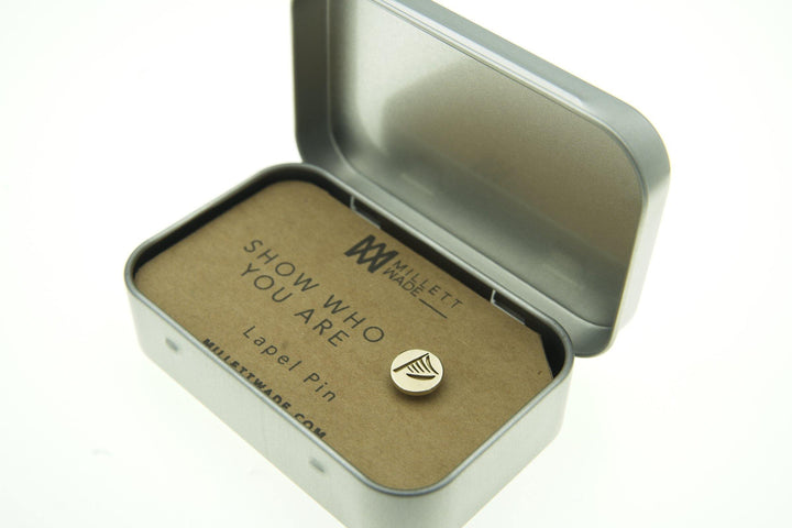 A small brass Harp Lapel Pin with a simple design is displayed inside an open metal tin. The hand-finished brass item is mounted on a piece of cardboard that reads "SHOW WHO YOU ARE." The tin and pin are from Millett Wade, as indicated by the branding on the packaging.