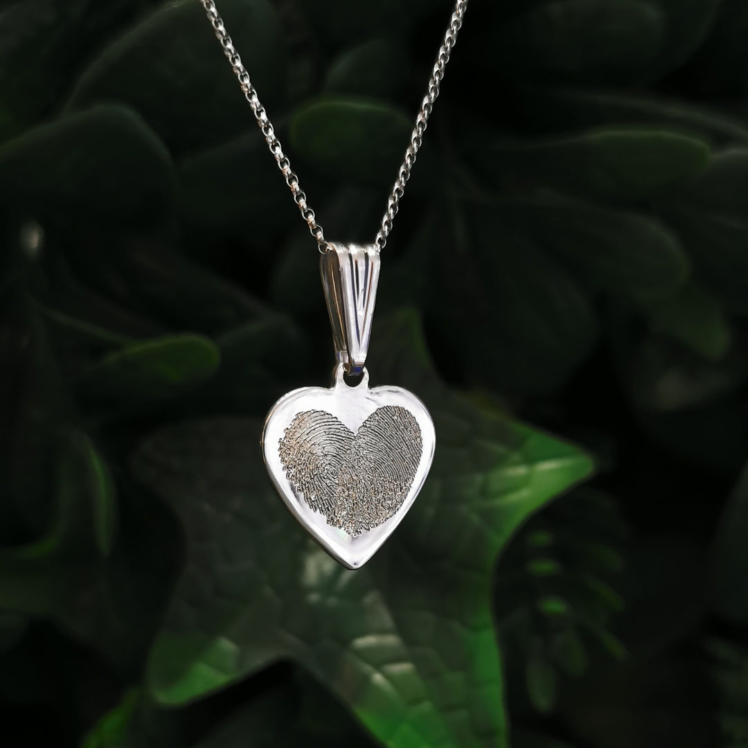 The Engraved Fingerprint Pendant by Personalised Charms features a silver heart-shaped design on a delicate chain, beautifully showcased against soft-focus greenery.