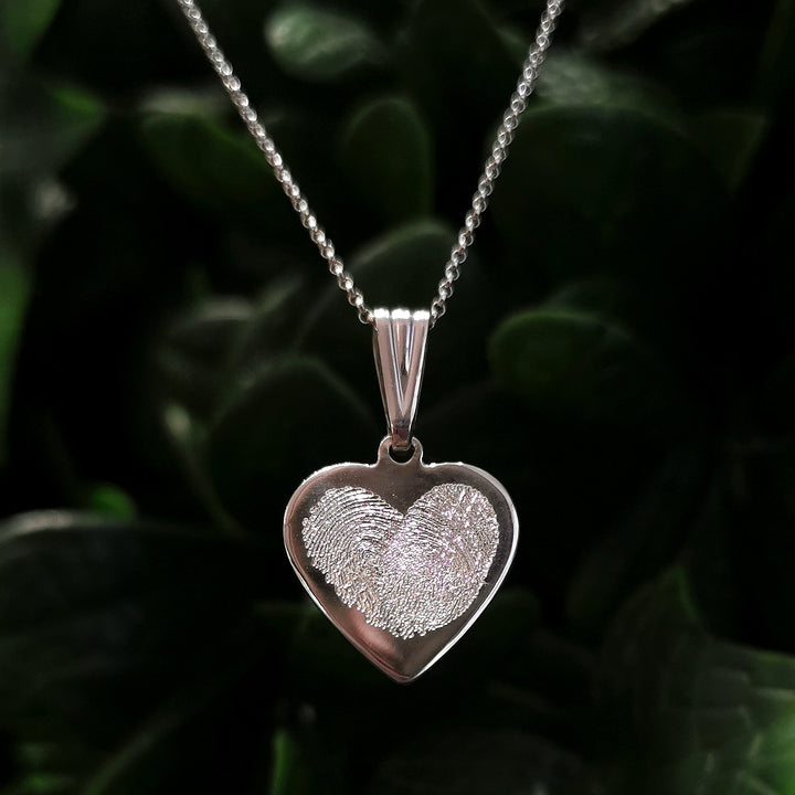 The Personalised Charms Engraved Fingerprint Pendant is a heart-shaped silver necklace with a delicately crafted fingerprint pendant, set against a dark green leafy background.