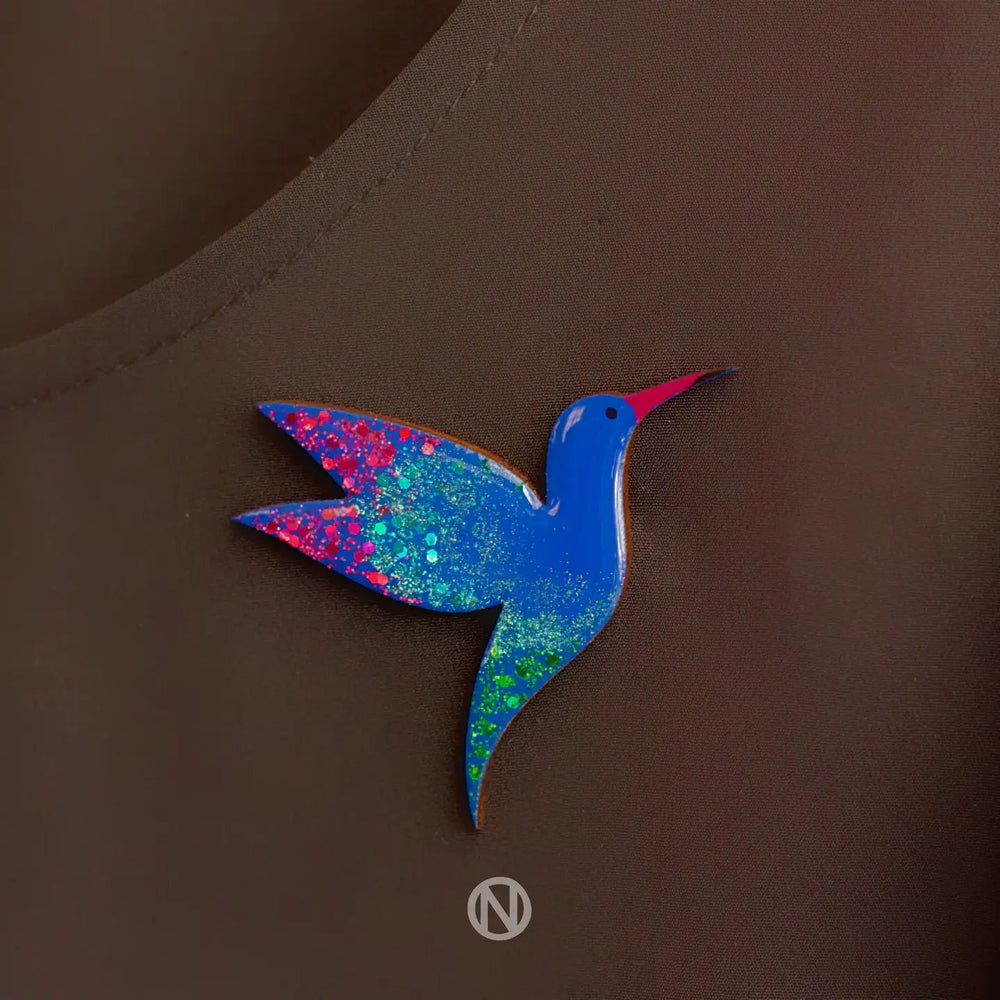 A brightly colored Naoi Hummingbird Pin Brooch is attached to a dark fabric, possibly a piece of clothing. The bird features blue, pink, green, and red specks, creating a vibrant, glittery effect reminiscent of hand-painted wooden ornaments.
