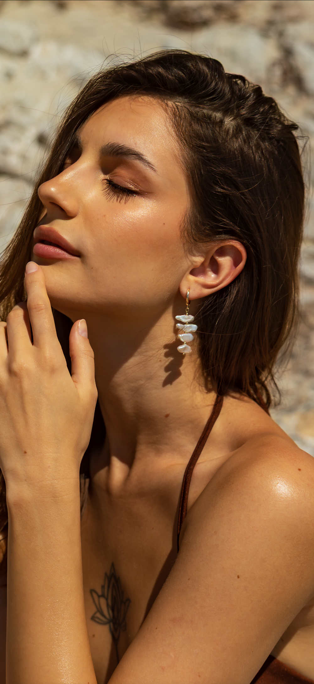 A woman with long brown hair poses outdoors, eyes closed and touching her chin. She wears a brown top, a lotus flower chest tattoo, and Kybalion Jewellery's gold plated Perla Sol Earrings. Sunlight highlights her glowing skin.