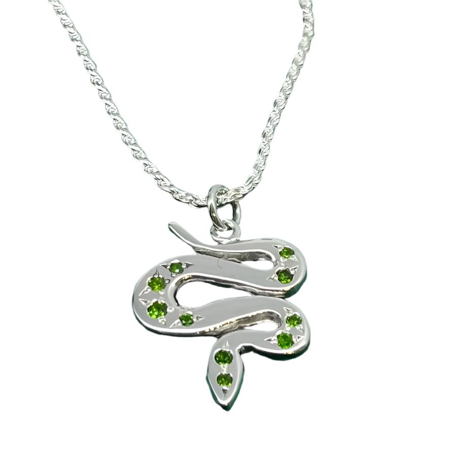 A Sterling Silver & Chrome Diopside Serpent Pendant by Cosmic Boulevard lies on a green fabric. The snake is curved into an "S" shape and is adorned with small green gemstones along its body, adding a touch of elegance and color to the piece. The chain is a simple twisted design.