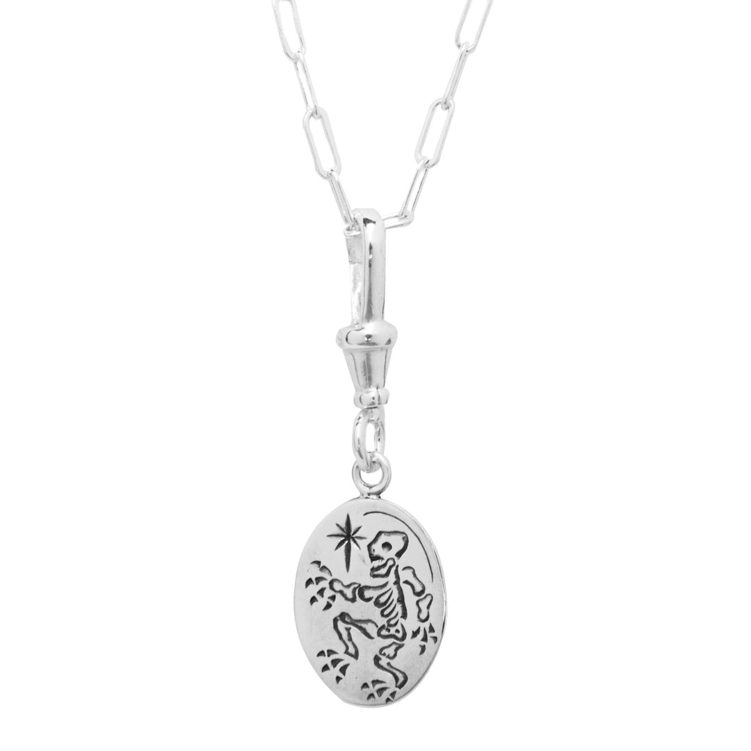 A sterling silver necklace with an elongated, rectangular link chain showcases a hand engraved pendant. This Cosmic Boulevard Memento Mori Silver Pendant features a skeleton dancing among stars and dots on an oval charm.