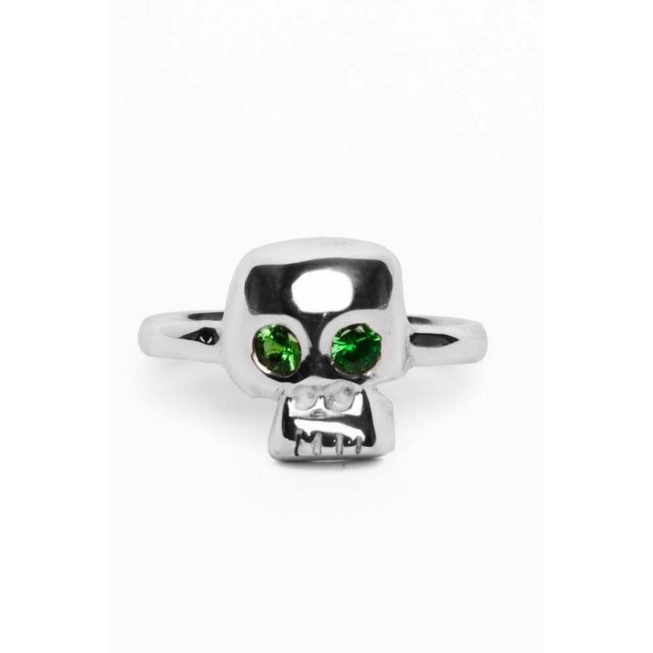 A **Silver Memento Mori Skull Ring** featuring a memento mori skull design with two bright green gemstone eyes. The sterling silver skull has a shiny, reflective surface and is centered on a polished, simple silver band, epitomizing the finest in handmade jewellery by **Cosmic Boulevard**.