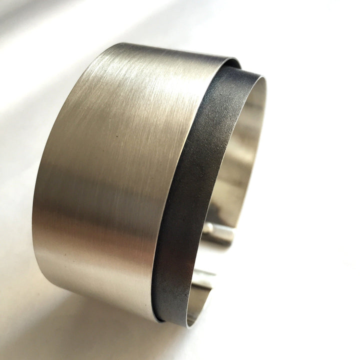 A sleek, modern stainless steel cuff bracelet with a brushed metal finish, part of the Bilbao Bracelet collection by MarCucurella. The band is wide and slightly open at the bottom, allowing for easy wear. The minimalist design features a smooth, clean surface, making this handmade jewelry a versatile accessory.