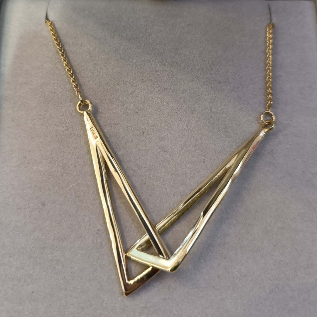 The Miriam Wade Le Cheile Pendant Large 9ct Yellow Gold features a unique geometric pendant with two overlapping open triangles forming an intricate V-shape. The handmade T-bar catch secures the fine gold chain, all displayed elegantly on a grey background.