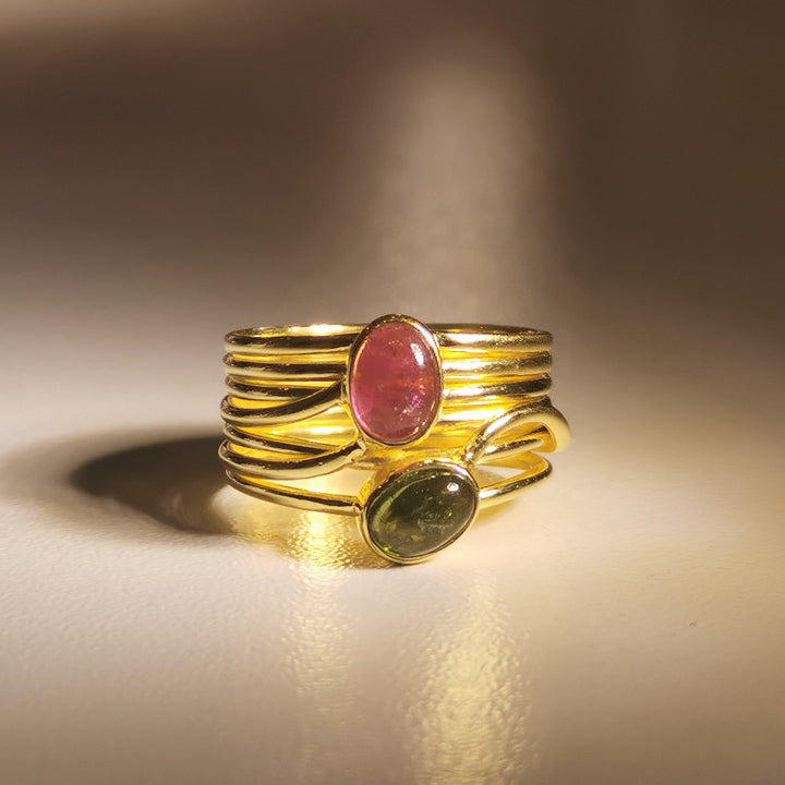 A Green and Pink Tourmaline in a Multi Band Gold Ring from Watermelon tropical is shown under soft lighting. It features two oval tourmalines: one pink and one green. The ring's handmade jewellery design intertwines the bands around the stones, creating an elegant and contemporary look.