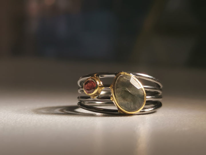 Close-up of a handmade jewellery ring with three bands merging into a single base. The center band holds an oval-shaped, reflective Labradorite stone framed in gold. Adjacent is a smaller, round, reddish-orange stone also set in gold. The Watermelon Tropical Black Rhodium and Gold Plated Ring with Labradorite and Garnet rests on a smooth surface with blurred background.