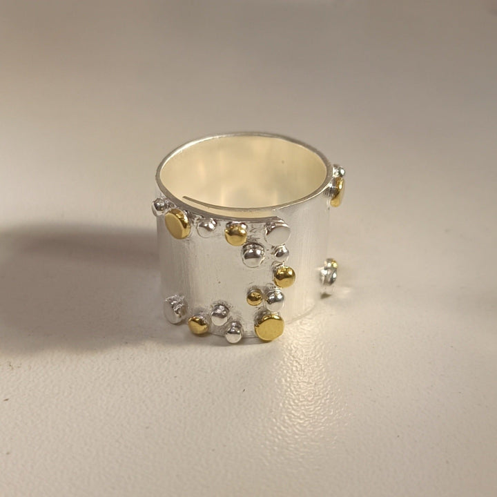 A wide, sterling silver ring with a matte finish, embellished with small, randomly placed round gold and silver beads, sits on a light-colored surface. The Iona Silver & Gold Ring by The Collective Dublin has a contemporary, artistic design.