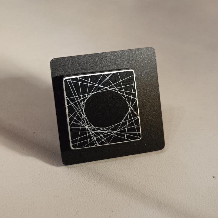 A black square object with a smaller square panel in the center. The central panel features a circular design created by intersecting hand-drawn lines, forming a web-like pattern. Crafted from high-end Acrylic, the Frey Ring by Alex + Svet stands on a light-colored surface against a plain background.