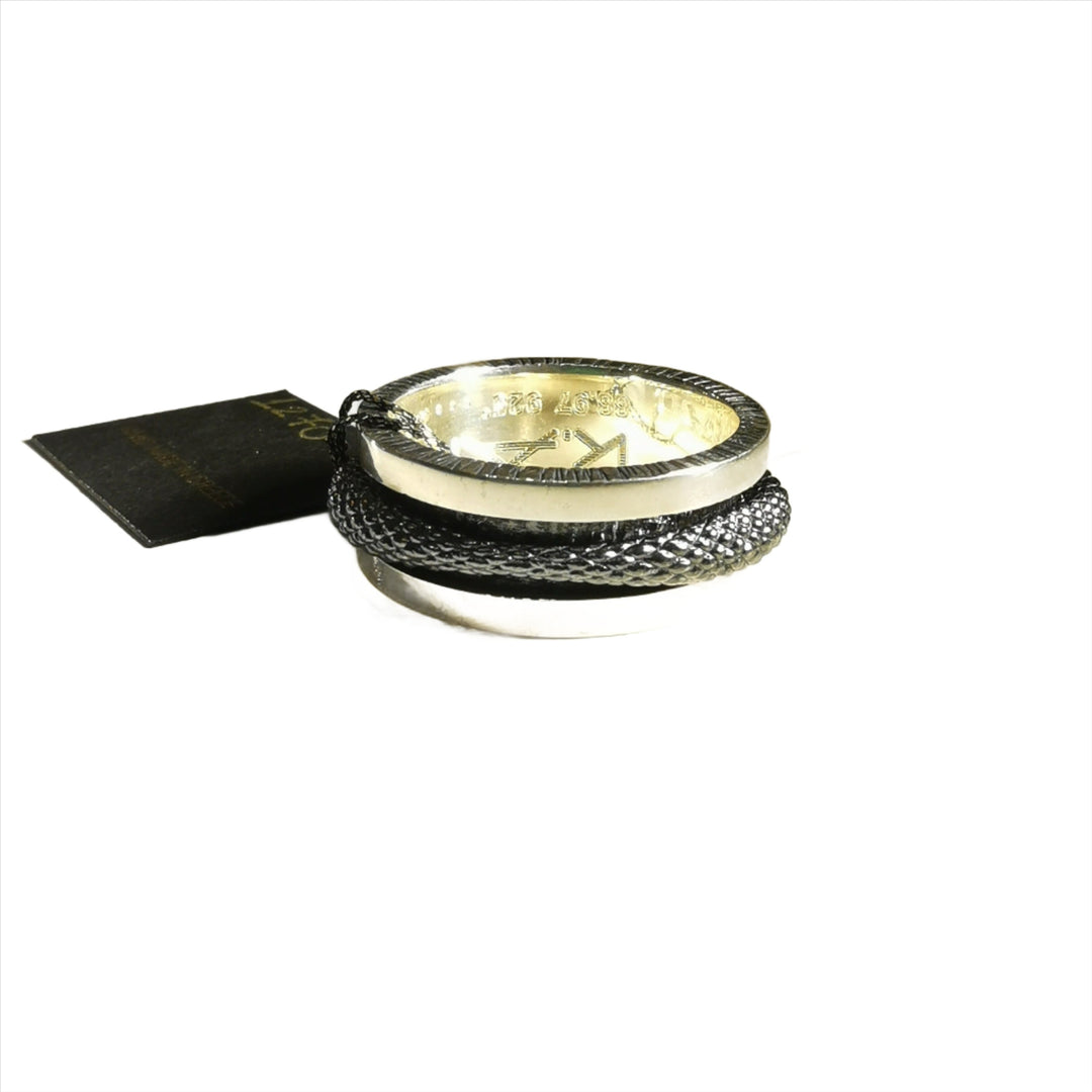 A Scaled Texture Silver Spinner Ring from K.And with an engraved design is wrapped with a dark, textured cord. A black tag with gold lettering is attached to the cord. The ring and cord are set against a plain white background.