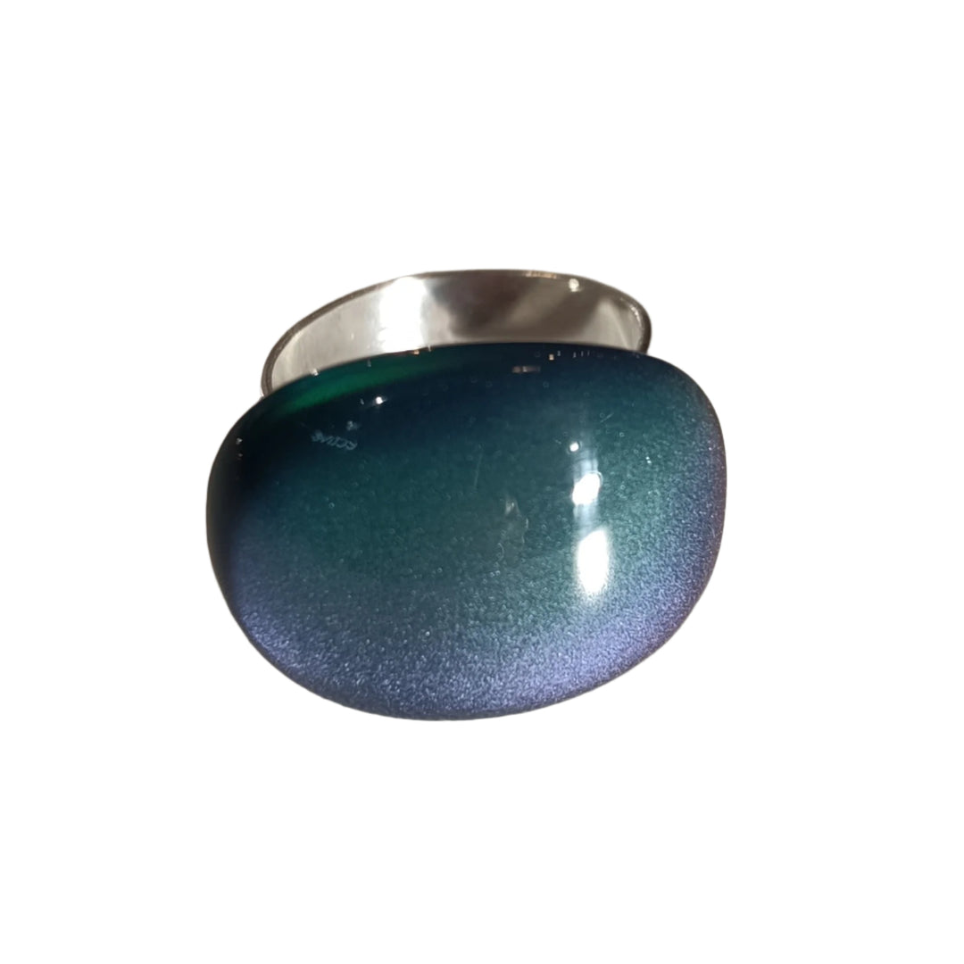 A MX.design Acrylic Blue/Green Ring featuring an oval-shaped, polished stone that transitions in color from teal to dark blue, reflecting light to create a gradient effect. The ring lies on a textured white surface, with soft shadows creating depth.