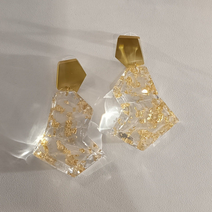 A pair of Acrylic Gold & Clear Earrings from MX.design lies on a white surface. Each earring consists of a beige, polygonal stud connected to a transparent, irregular-shaped dangle embedded with gold flakes. The design combines a rustic and modern aesthetic.