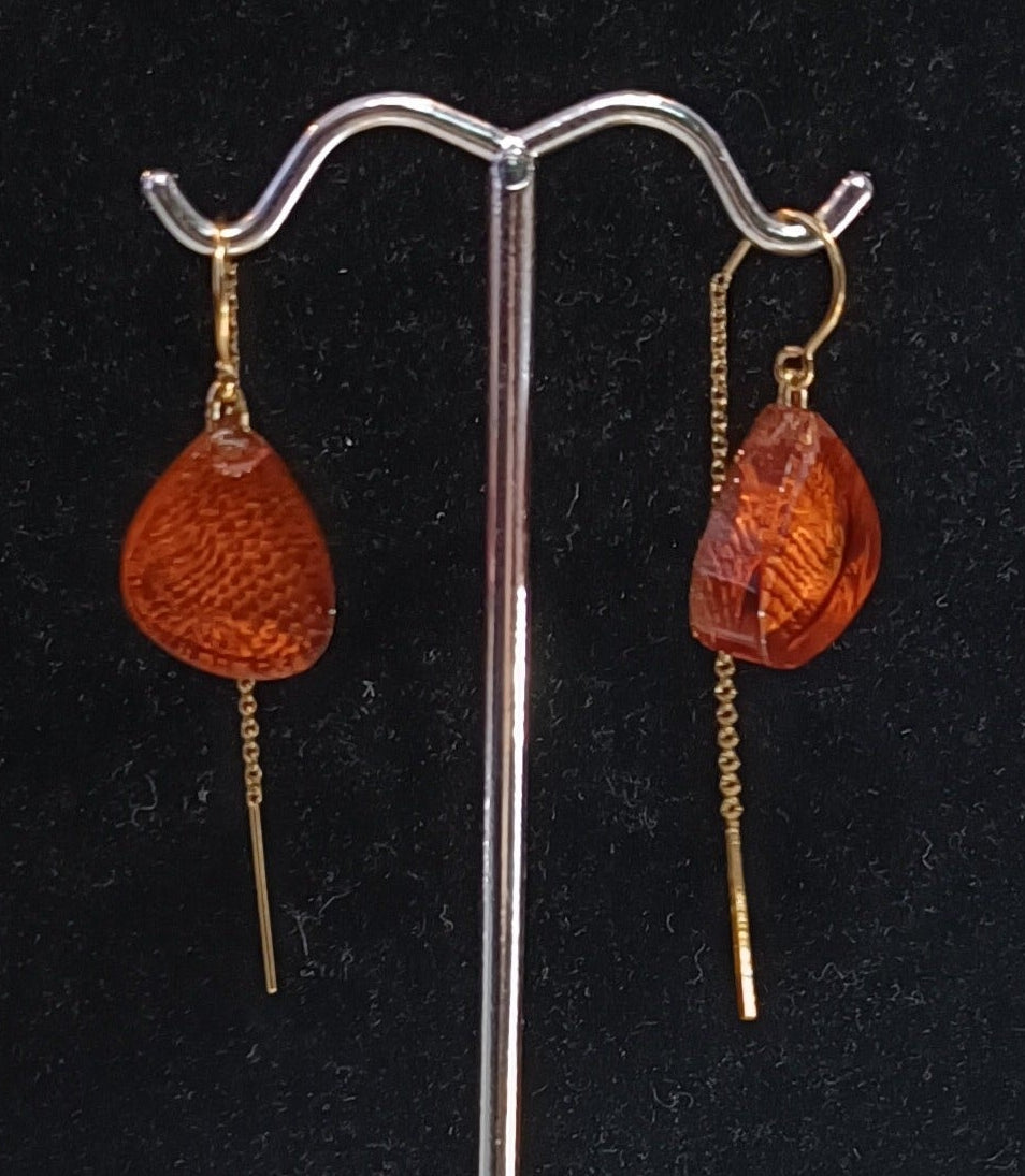 A pair of orange, teardrop-shaped Acrylic Rust Earrings with Chain Back Gold by MX.design, hanging from a silver display stand against a black background. One earring features a dangling chain element, while the other has a small, orange bead above the teardrop.