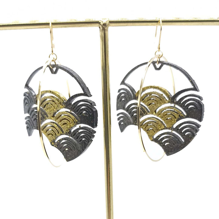 A pair of Black Japanese Wave Earrings by Tochu-De is displayed on a metallic stand. They have intricate wave-like patterns in black and gold, with an outer gold hoop. Crafted from 3D printed nylon, the design features a detailed, artistic style blending modern and traditional elements. For jewellery care, use a soft dry cloth.