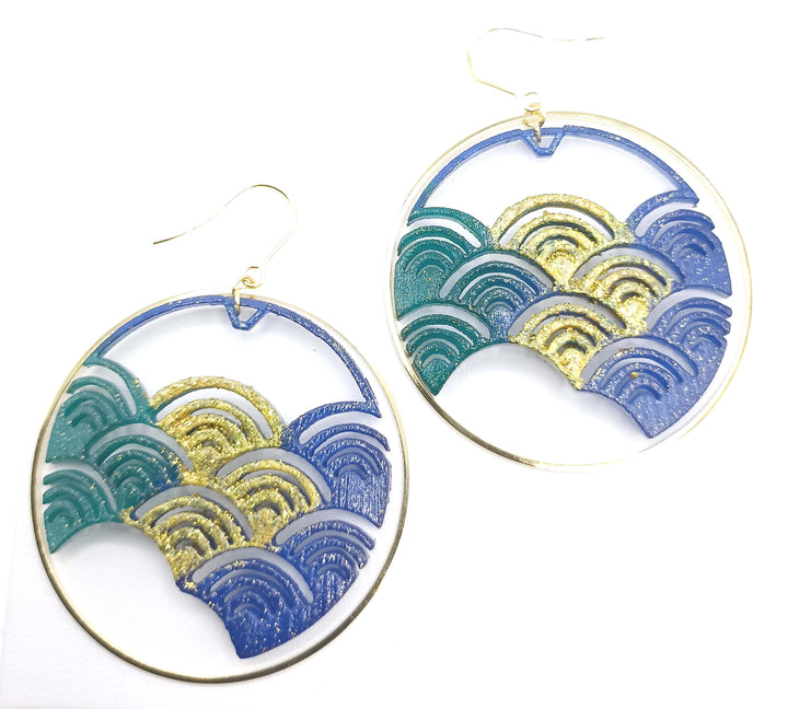 A pair of Blue Japanese Wave Earrings, 3D printed in nylon and featuring an intricate wave pattern in shades of blue, teal, and gold. The design by Tochu-De includes glittery accents, and the earrings have thin, gold-colored hooks for fastening. The background is plain white.