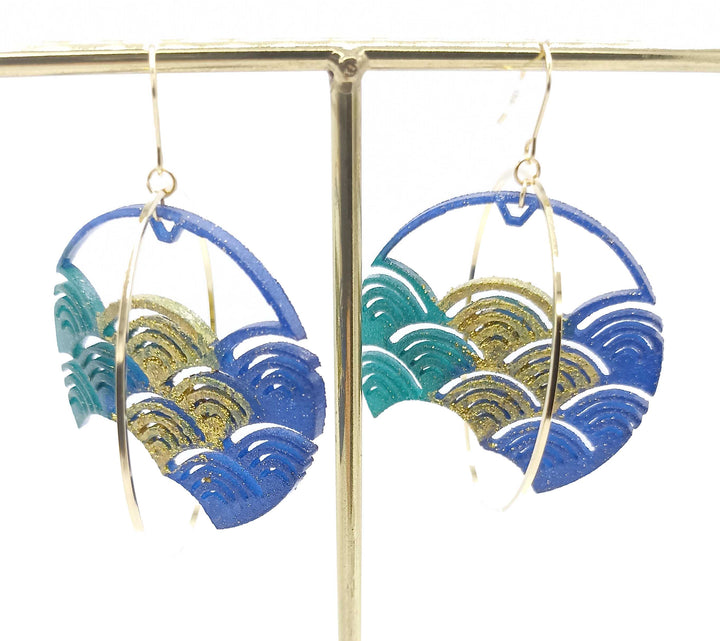 A pair of dangling earrings with wave designs in shades of blue and gold glitter. These 3D-printed nylon Blue Japanese Wave Earrings by Tochu-De have circular cut-outs and are suspended from gold hooks, hanging on a metallic display stand against a white background. Remember jewellery care to keep them sparkling.