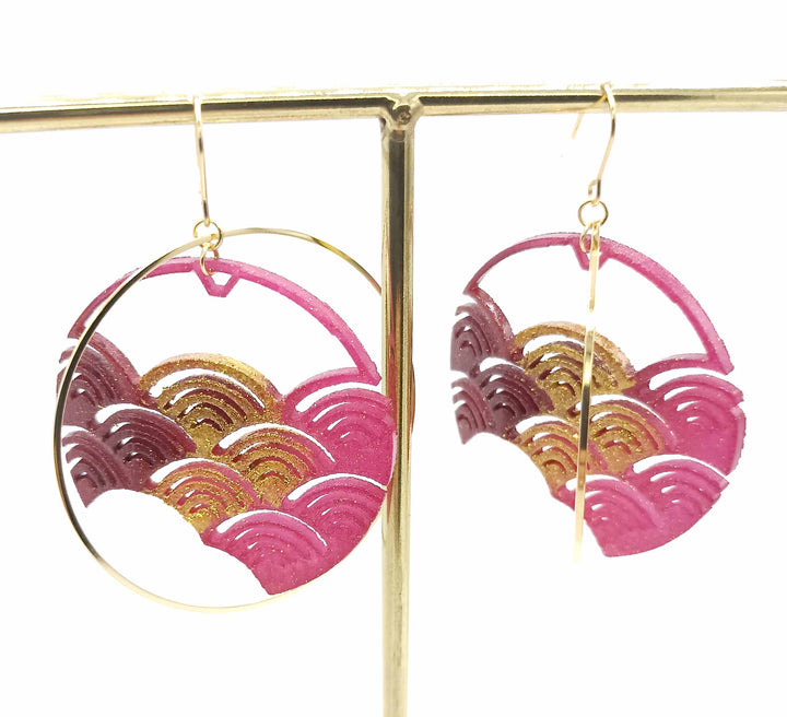 Circular dangle earrings featuring intricate pink and gold wave patterns suspended from gold hooks. The design includes a combination of metallic and vibrant colors, giving the powder-coated jewellery an elegant and artistic look. The Fuchsia Japanese Wave Earrings by Tochu-De hang from a gold display stand.