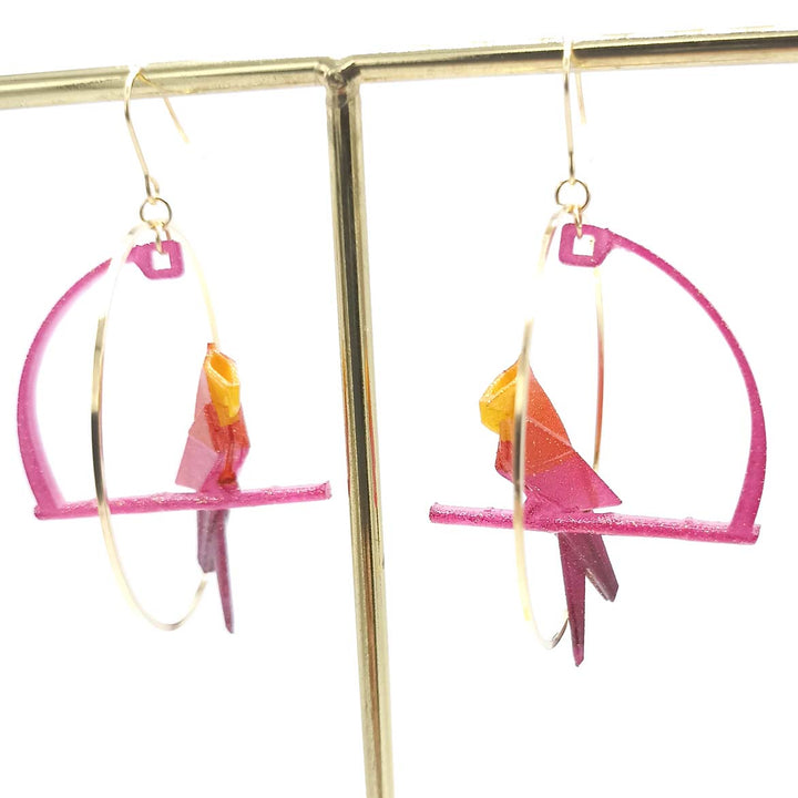 A pair of colorful Fuchsia Origami Parrot Earrings from Tochu-De are displayed on a jewelry stand. The earrings, made from 3D printed nylon, feature pink hoops with bright pink and orange parrot figures perched in the center. The design is vibrant and playful.