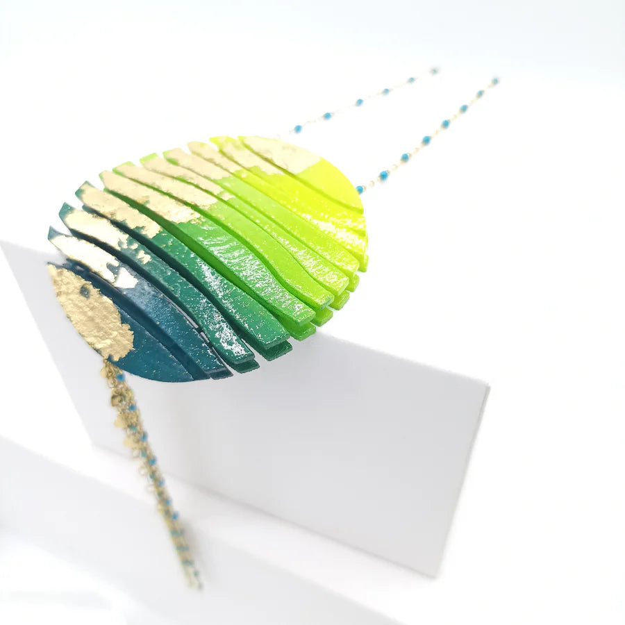 The Tochu-De Long Circle Wave Necklace in Petrol & Citrus is a round, layered statement piece with blue, green, and yellow gradients, gold accents, and small blue beads on the chain. Displayed on a white background.