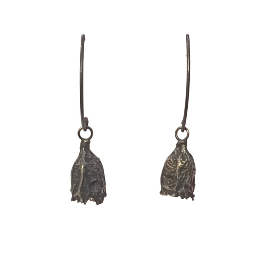 A pair of oxidised sterling silver Husk Drop Earrings by Abbie Dixon displayed on a white stand. The earrings dangle on hooks and feature intricate, slightly rugged drop-shaped designs, giving them a rustic and handcrafted appearance.