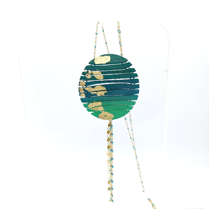 A Long Circle Wave Necklace in Petrol & Emerald by Tochu-De hangs from a beaded chain. The pendant has a layered, textured design and the chain, measuring 28", is adorned with small gold and turquoise beads for an elegant appearance. Crafted from 3D printed nylon, the jewellery comes on a plain white backdrop.