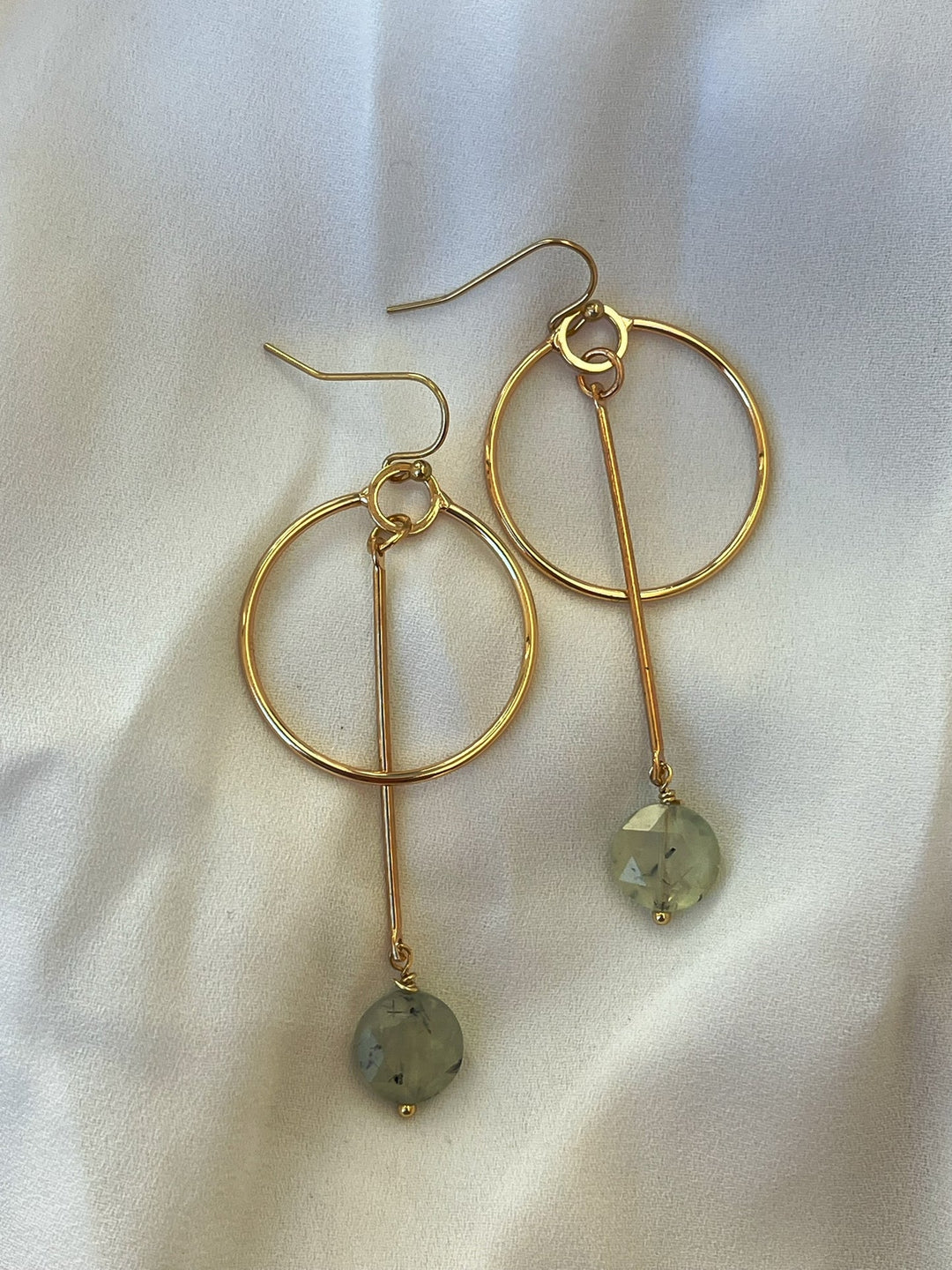 The Kea Earrings with Prehnite Drop by Kybalion Jewellery feature gold-plated bars and faceted light green prehnite beads, showcased on a soft, light-colored fabric background.