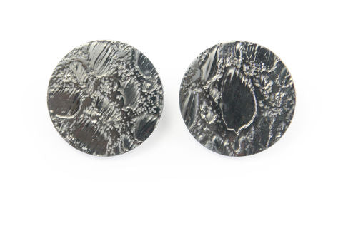 Two circular silver Etched 'Skin' Stud Earrings by Sara Gunn with a rugged, textured surface. The design features various raised and recessed areas, creating a metallic, rough look. They resemble finely crafted textured pieces and are placed against a plain white background.