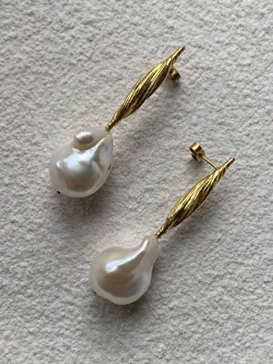 The Inner Island Gathered Forms Earrings feature gold vermeil studs with elongated, leaf-like designs and large, irregular white baroque pearls, shown on a textured light surface.