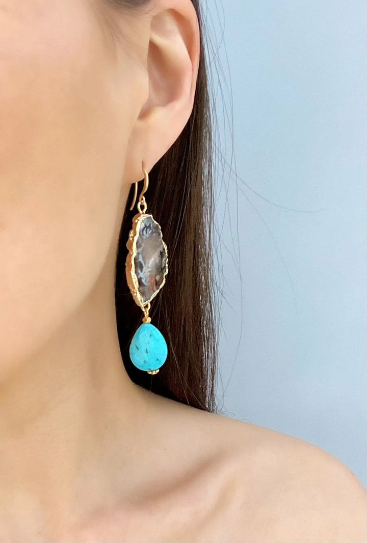 A close-up of a woman's ear wearing the Aria V Cameo Earrings. The earring features an irregularly shaped, textured gold and clear stone on top with a turquoise teardrop hanging below. The background is plain and light-colored.