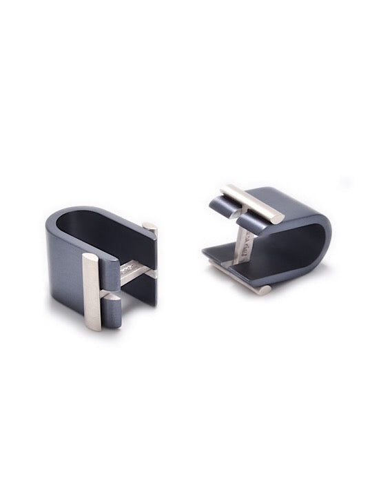 Two Affinor Wrap Around Cufflinks by Filip Vanas with a modern, sleek design made from sterling silver anodized aluminum. Each cufflink has a U-shaped structure with a small bar across the top to secure cables in place. The one on the left is standing upright, while the one on the right is lying on its side.