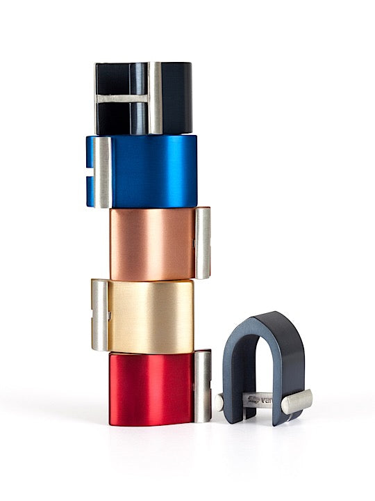 A stack of colorful, modern, cylindrical metal Ogive Wrap Around Cufflinks by Filip Vanas in various finishes, including blue, copper, gold, and red, beside an individual cufflink in black. Each clip has a sleek, minimalist design with a small cylindrical rod for fastening—an innovative concept akin to stylish cuff links.