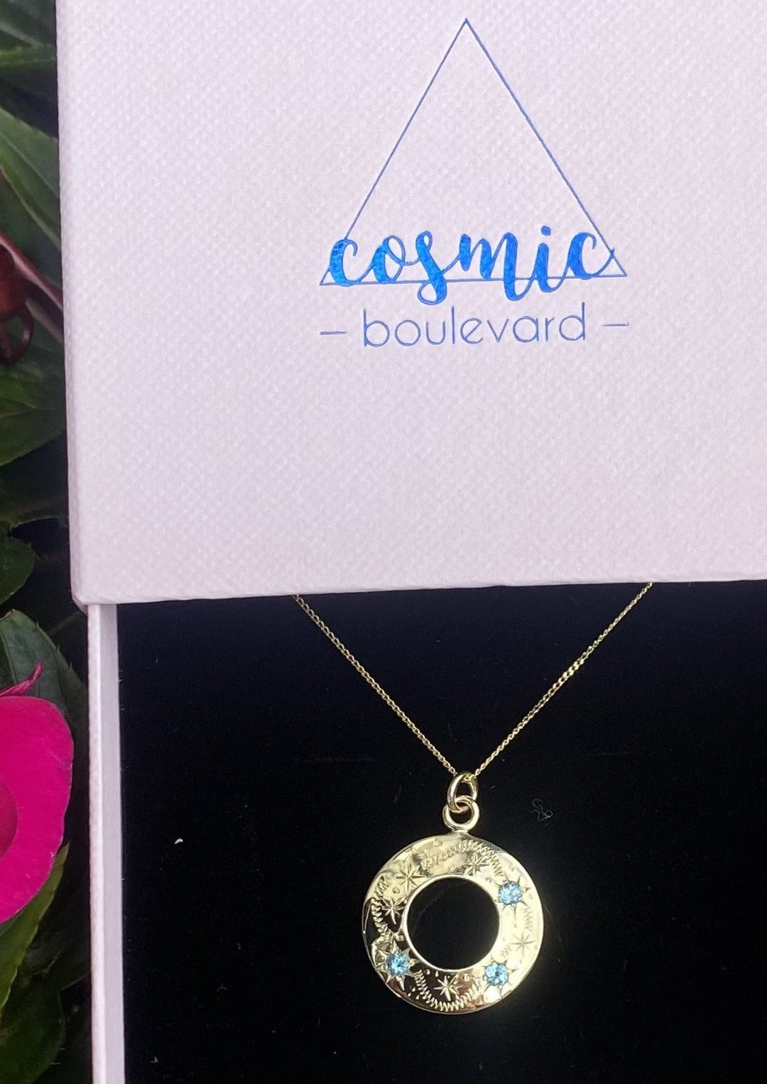 The "Something Blue" Shooting Star Pendant in gold, crafted by Cosmic Boulevard, is elegantly showcased in an open white jewelry box. The circular pendant, adorned with star designs and delicate blue topaz stones, catches the eye. The lid of the box features a triangular logo with the text "Cosmic Boulevard" in blue. In the background, pink flower petals add a dreamy touch ideal for sky lovers.