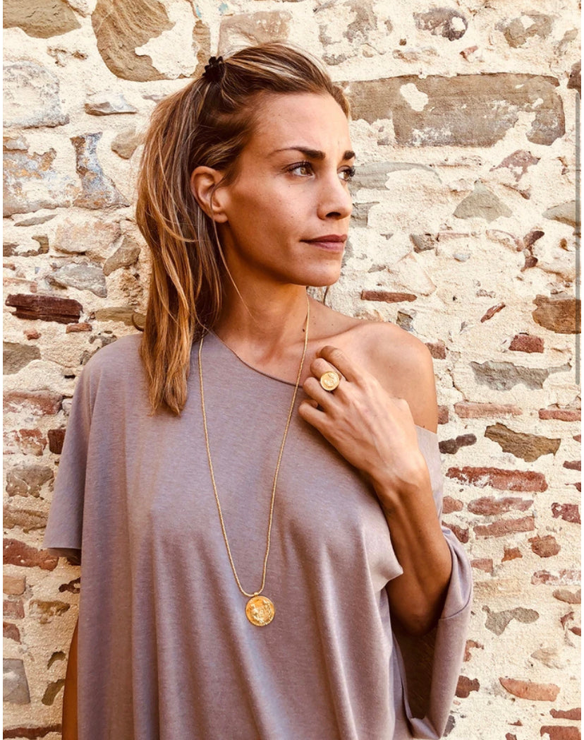 A woman with long, light brown hair is standing in front of a stone wall. She is wearing an off-shoulder taupe top and a Silver Gold Plated Jennifer Kinnear Grace O'Malley Medallion Small necklace. She also has a matching gold ring on her left hand and gazes thoughtfully to her left.