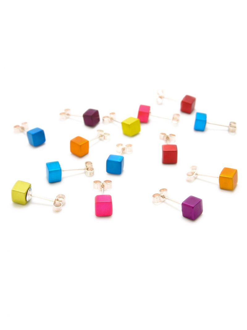 An assortment of colorful cube-shaped Small Cube Stud Earrings by Filip Vanas is scattered across a white background. Various vibrant colors, including red, blue, orange, pink, and purple, are featured. Each earring has an anodized aluminum stud and backing.