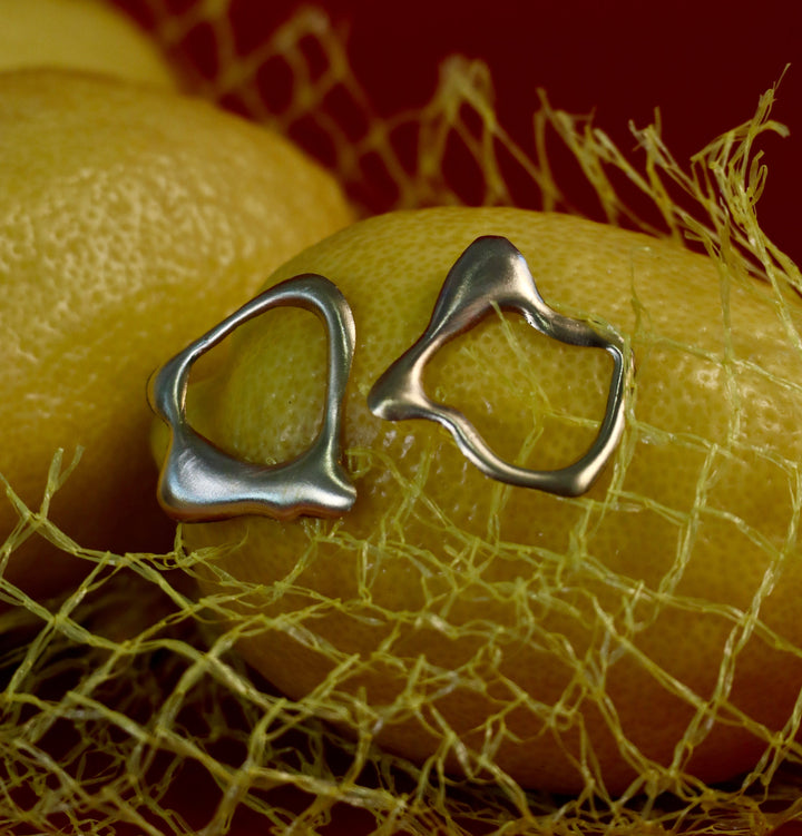 Molten Studs by Madeleine Sculptural Jewellery—abstract silver earrings with organic, irregular shapes—sit atop a yellow lemon in yellow netting, set against a vibrant red background.