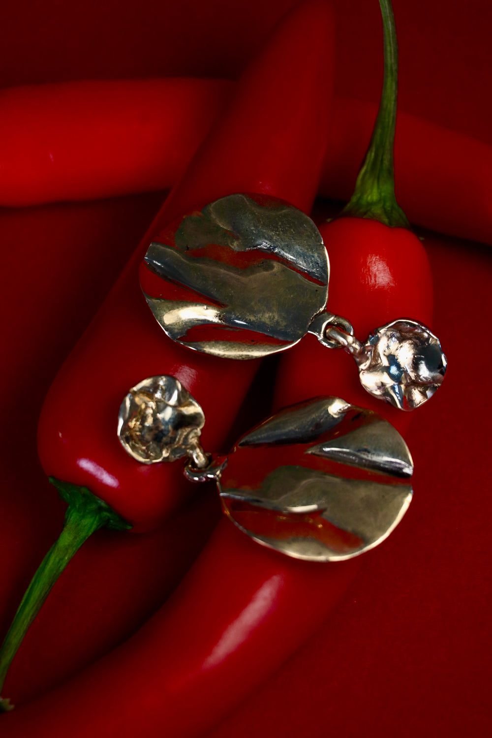 The Tide Drop Earrings by Madeleine Sculptural Jewellery—round and textured in silver—are shown atop red chili peppers against a matching background, their shiny, irregular surfaces reflecting light for a striking effect.